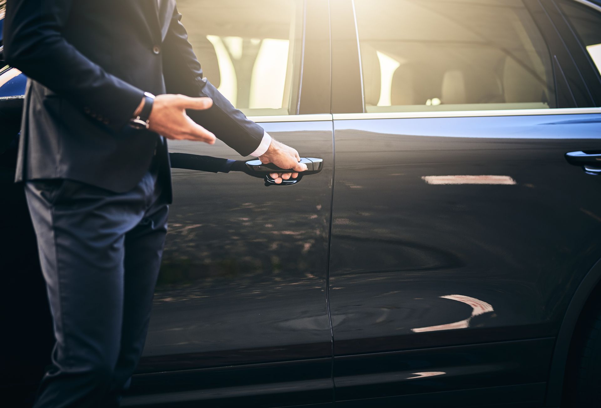 A person in a suit stands by a car, holding the door handle open to welcome a passenger.