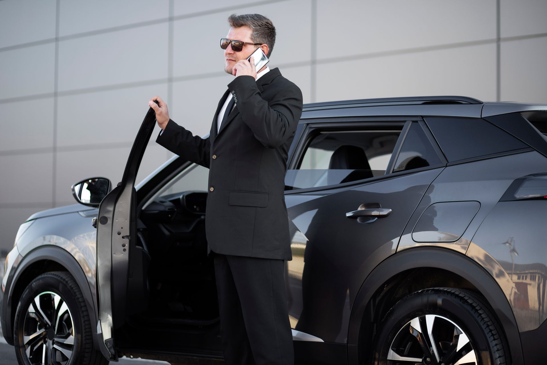 Man in a black suit and sunglasses stepping from a dark SUV outdoors, talking on a phone
