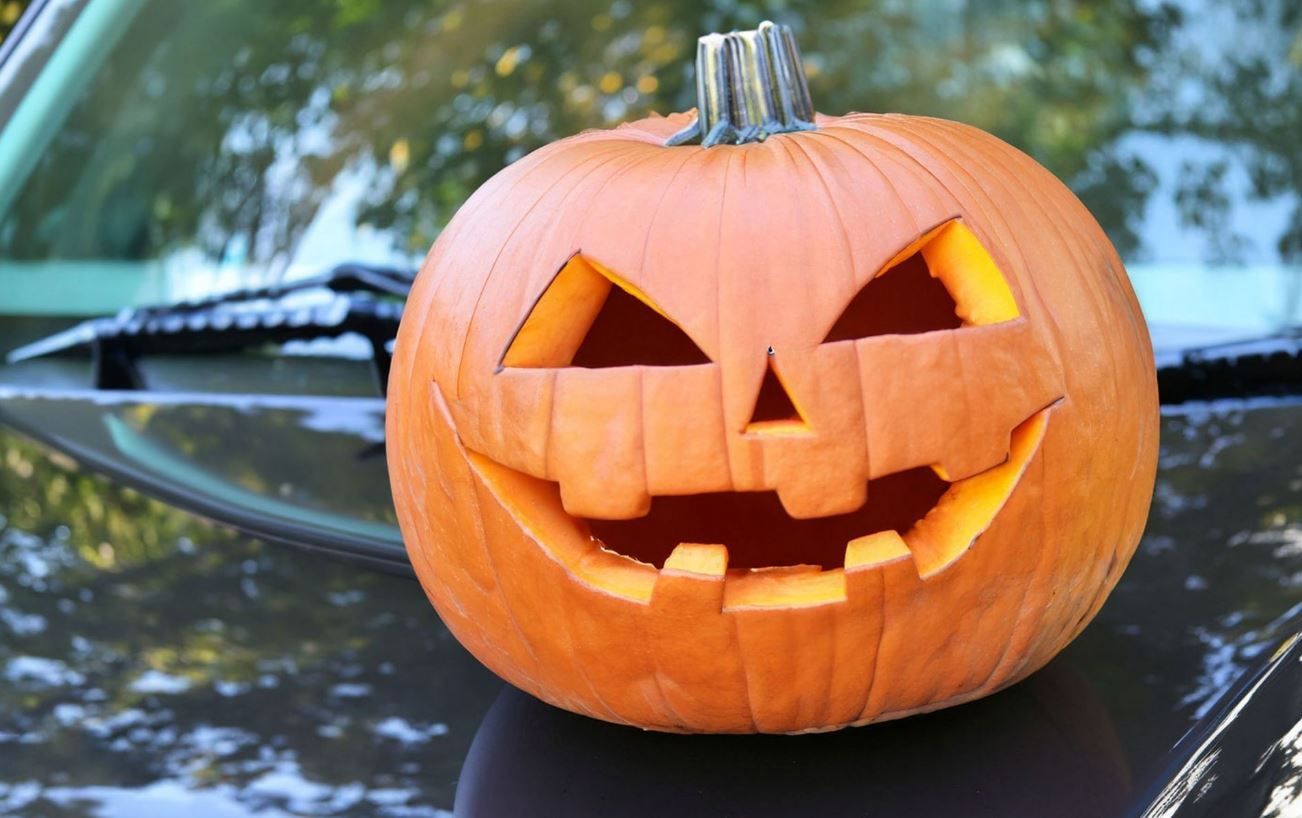 Carved orange jack-o'-lantern on a car hood with trees reflected in the glossy black surface