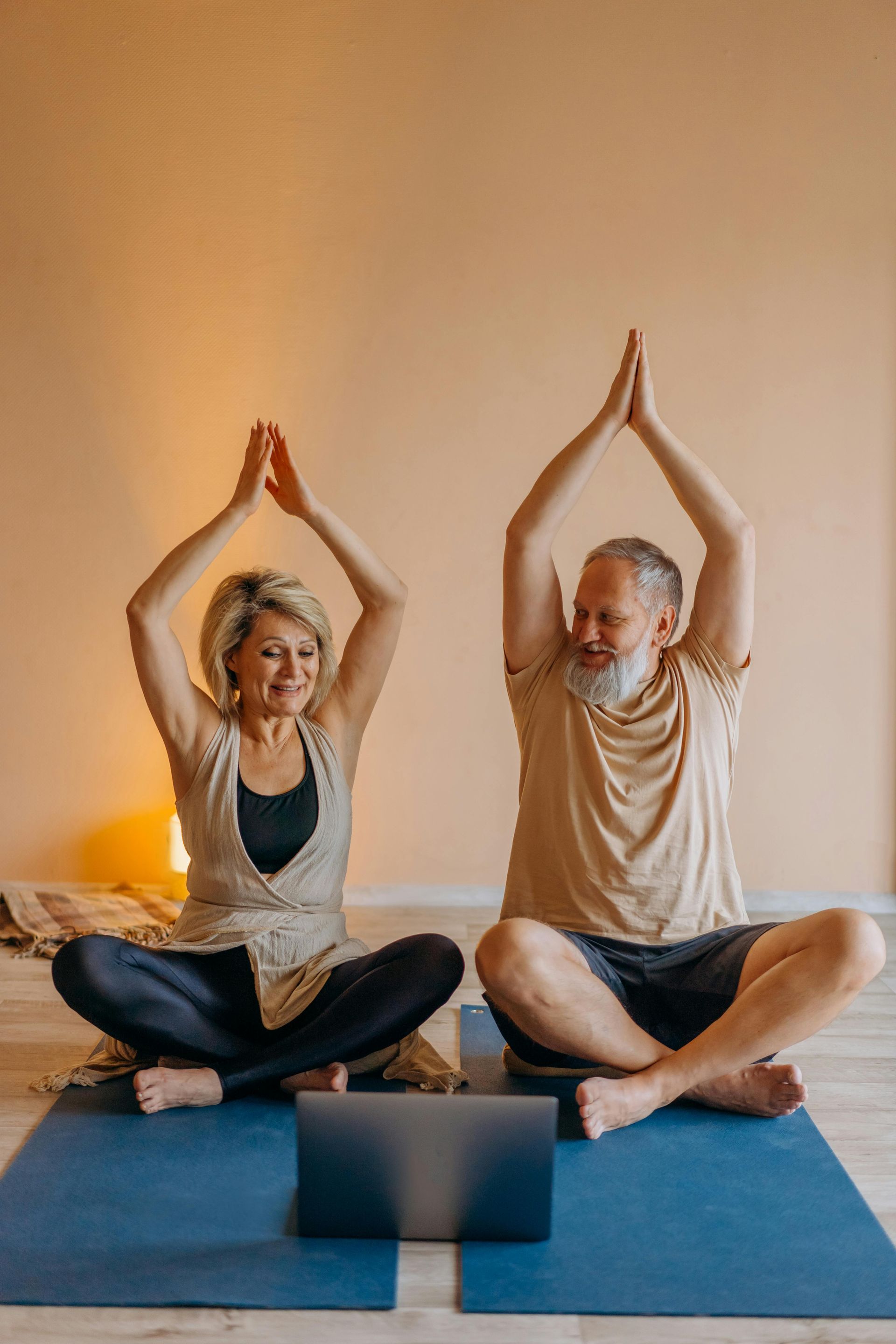 Elderly couple doing yoga together, sitting on mats in front of a laptop, arms raised in prayer pose. They smile in a warm-toned room.