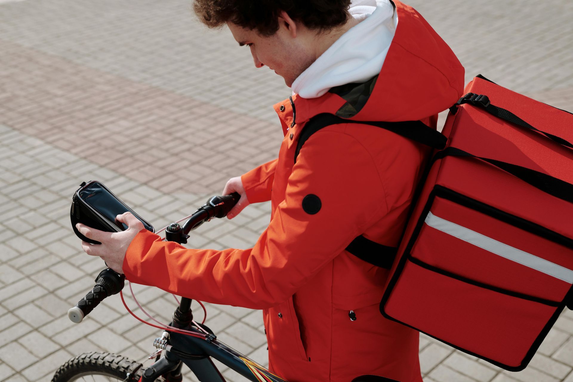 A person in a red jacket and backpack checks a phone mounted on their bicycle handlebars while outdoors.