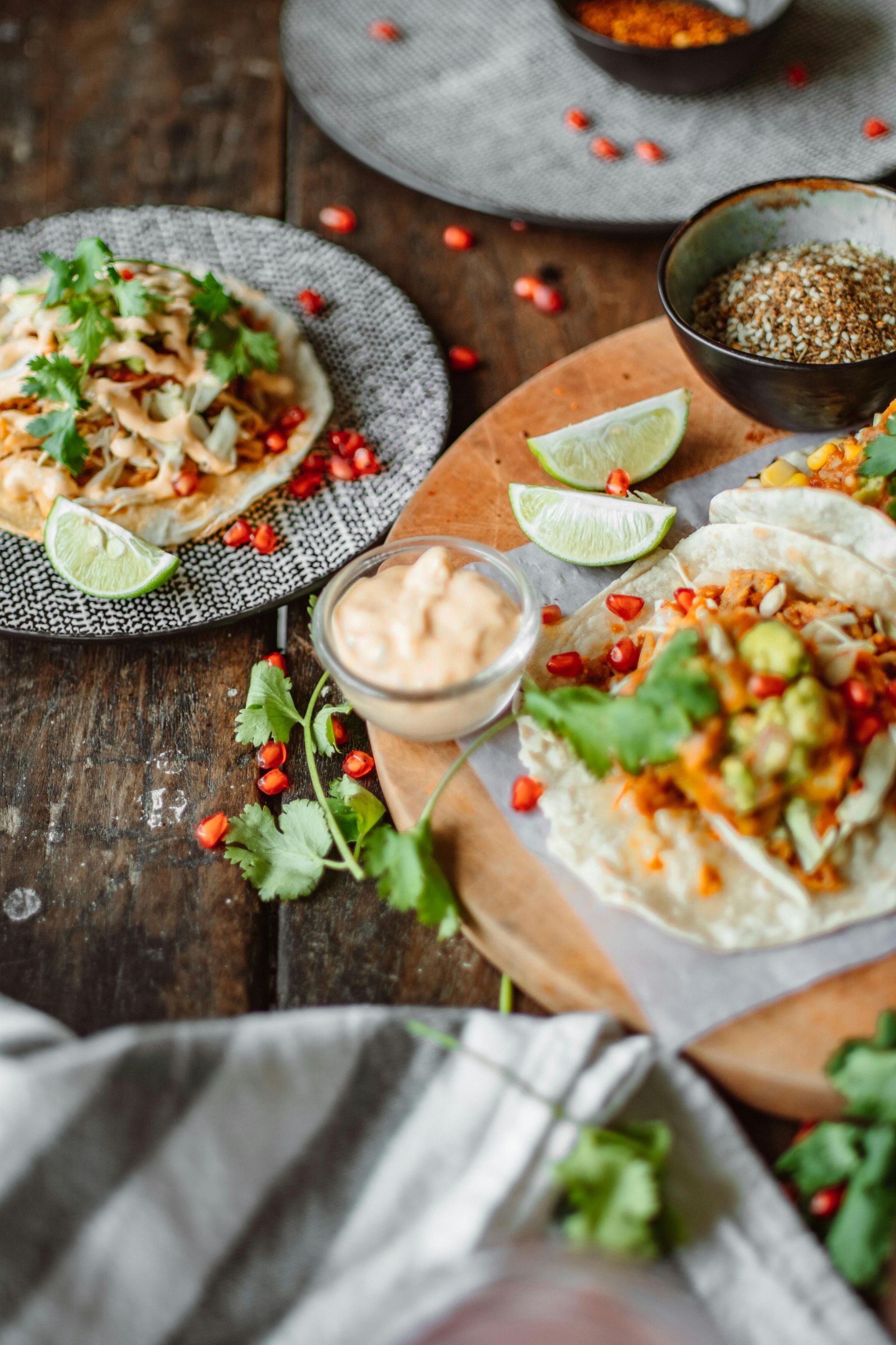 Tacos topped with cilantro, pomegranate seeds, and creamy sauce on plates and a wooden board with lime wedges and spices.