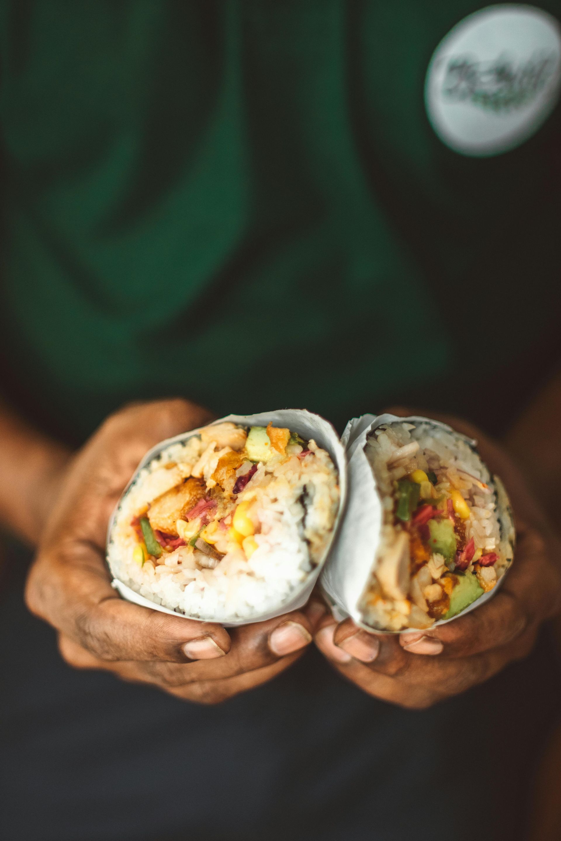 A person in a green uniform holds two halves of a burrito filled with rice, vegetables, and meat, wrapped in foil.