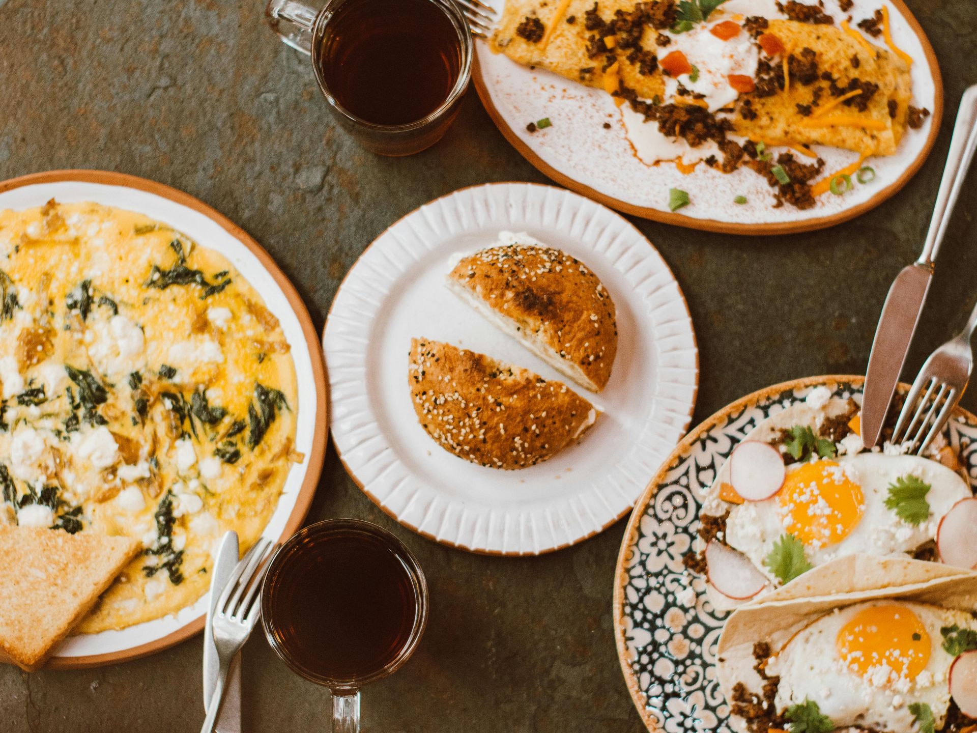 A top-down view of a rustic breakfast spread featuring omelets, eggs, a split bagel, and two mugs of coffee on a dark table.