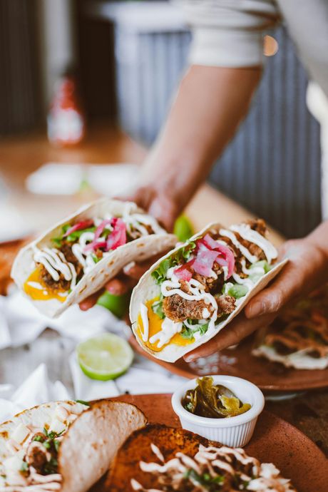 Hands hold two tacos topped with pickled red onions and creamy sauce, with more tacos and a small bowl of garnish nearby.