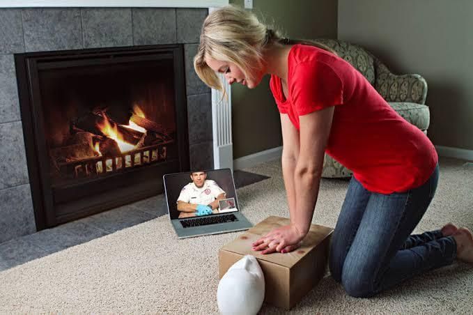 Woman kneeling by a fireplace, opening a cardboard box with a baby monitor showing an infant
