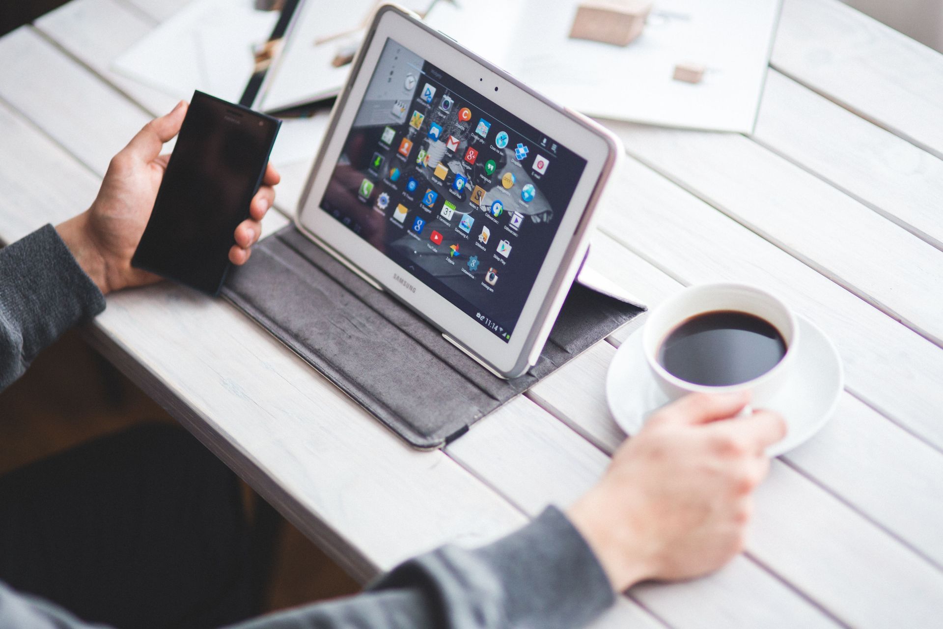 Hands holding a smartphone and coffee beside a tablet on a desk