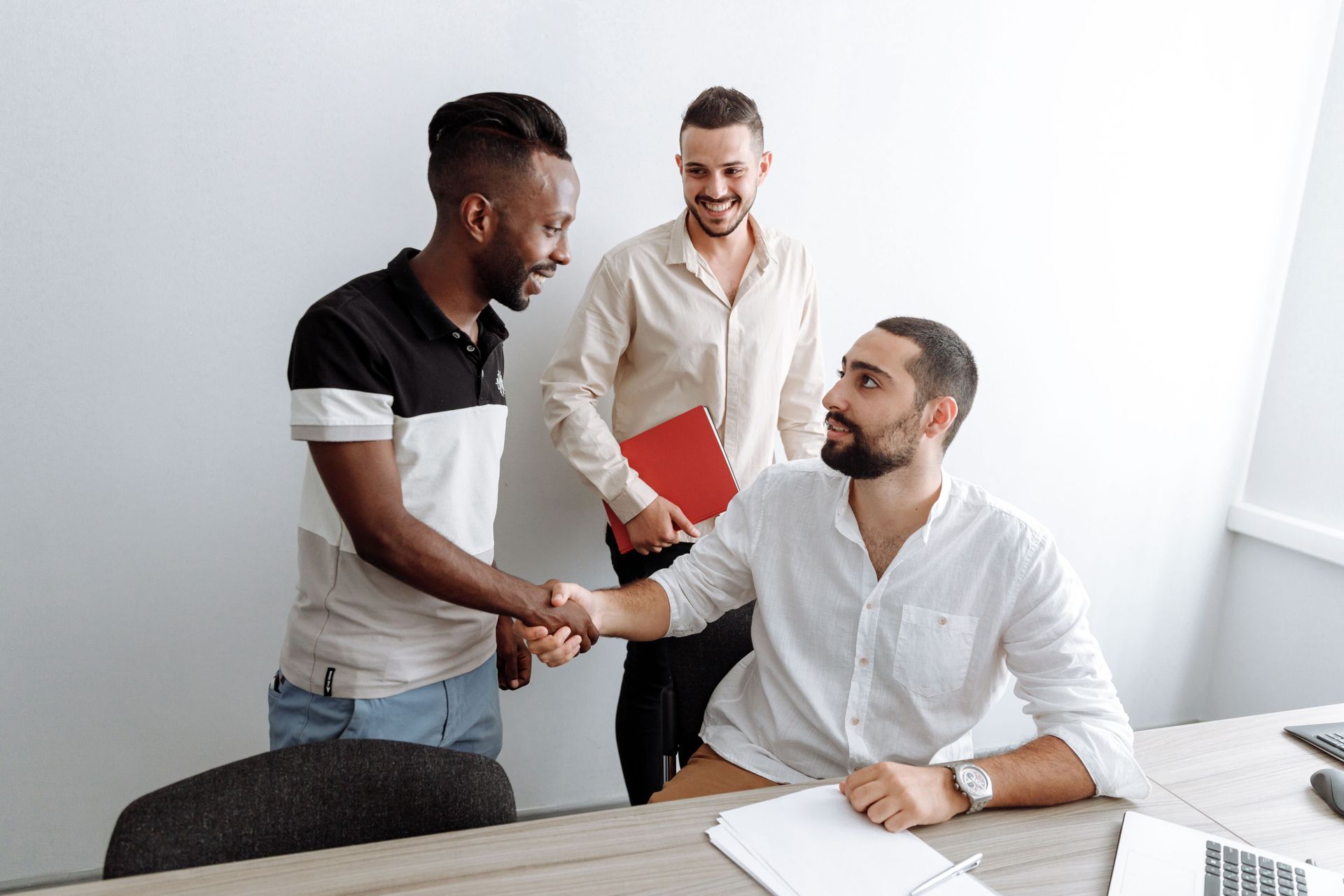 Three colleagues shaking hands in a bright office, one holding a red folder.