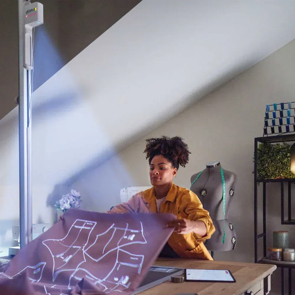 Person in a mustard shirt at a desk, holding a translucent sheet with white line drawings in a bright room.
