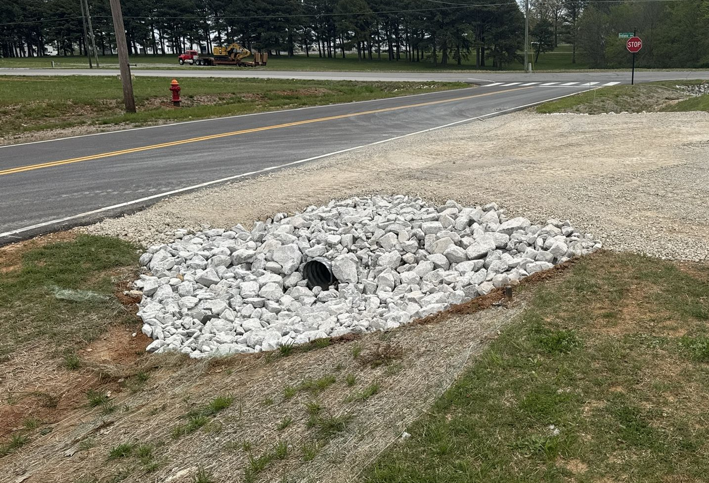 A culvert pipe embedded in a pile of gray riprap rocks at the edge of a paved road and gravel driveway.