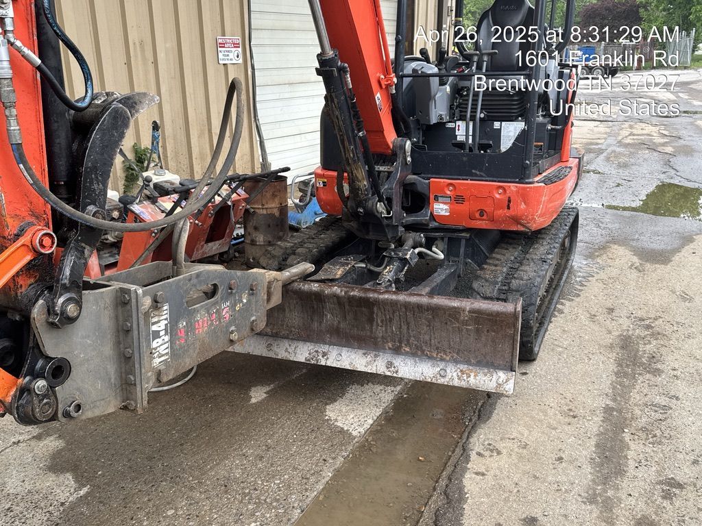 An orange mini excavator with a metal blade attachment parked on a paved lot.