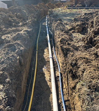 Trench with buried utility lines: yellow gas, white pipe, and black electrical cable. Soil fills the trench.