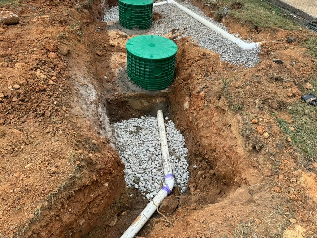 Two green drainage catch basins installed in a dirt trench with gravel and white PVC piping.