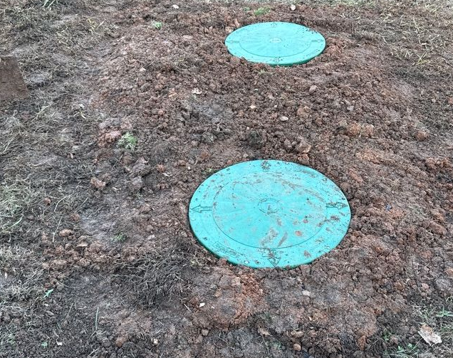 Two circular green plastic septic tank access covers set into a patch of brown soil.