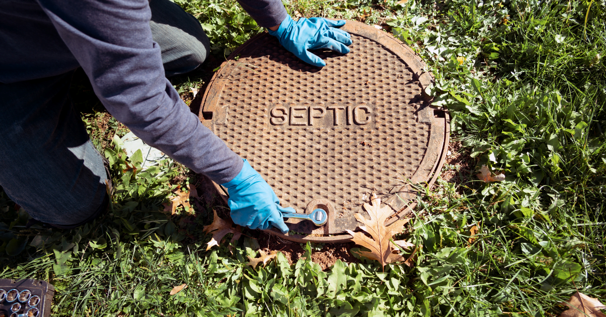 A person wearing blue gloves uses a tool to open a round, rusted metal cover marked 