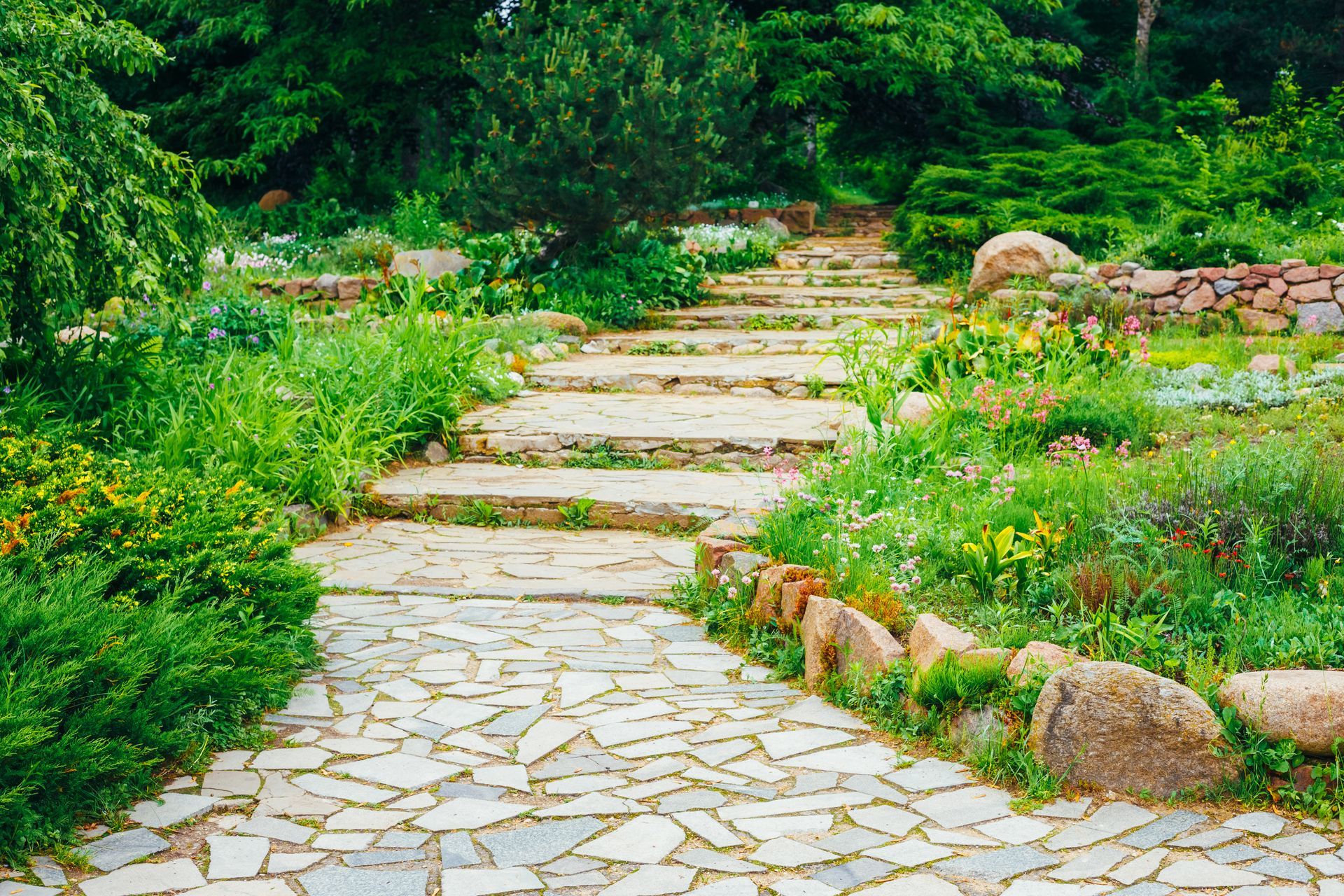 Stone path with steps ascends through a garden, lined with greenery and rocks.