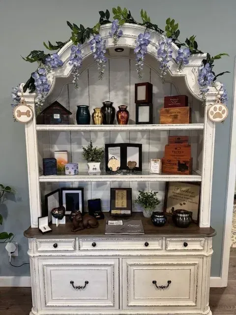 White hutch display with shelves, jars, plants, and purple flower garland; two drawers below.