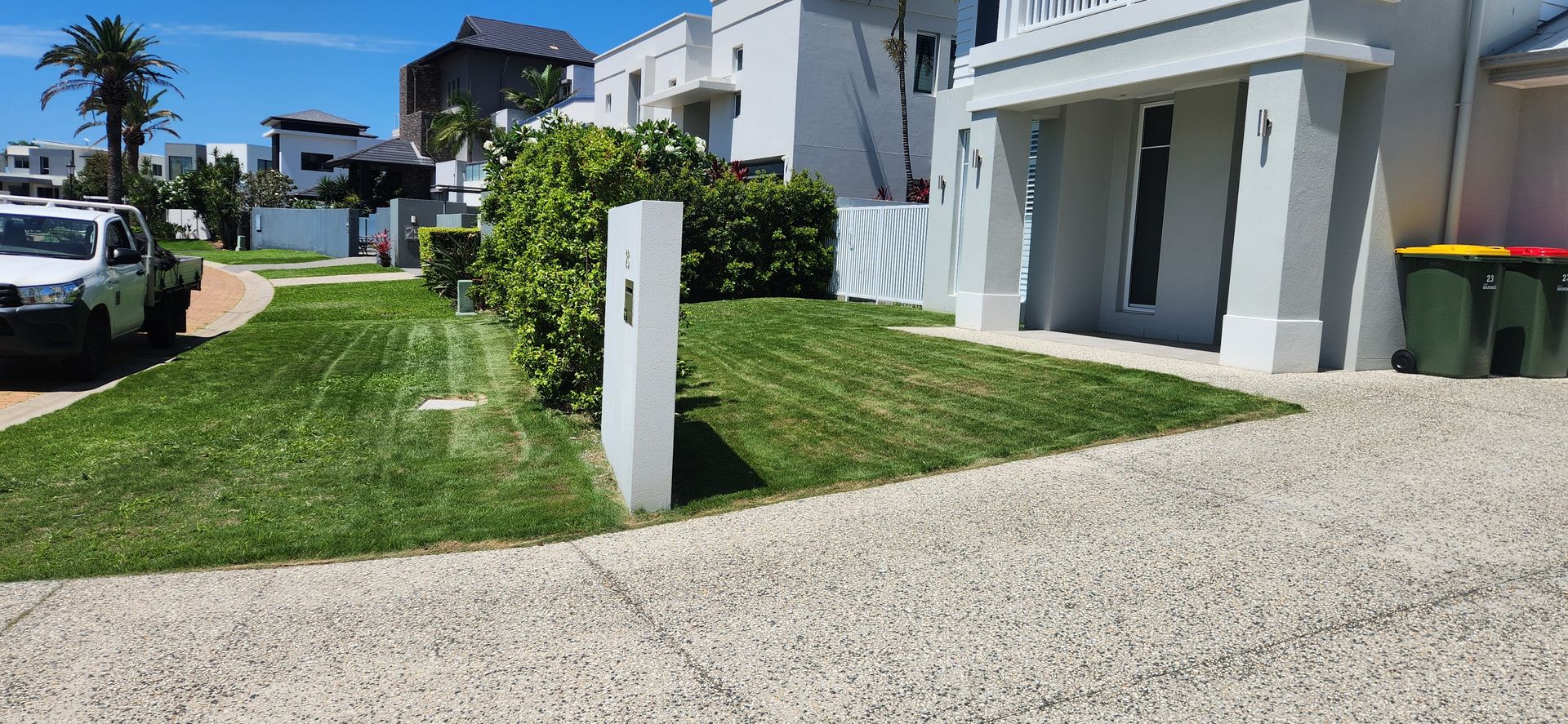 Sunny residential driveway beside white houses, green lawn, and parked cars under a clear blue sky