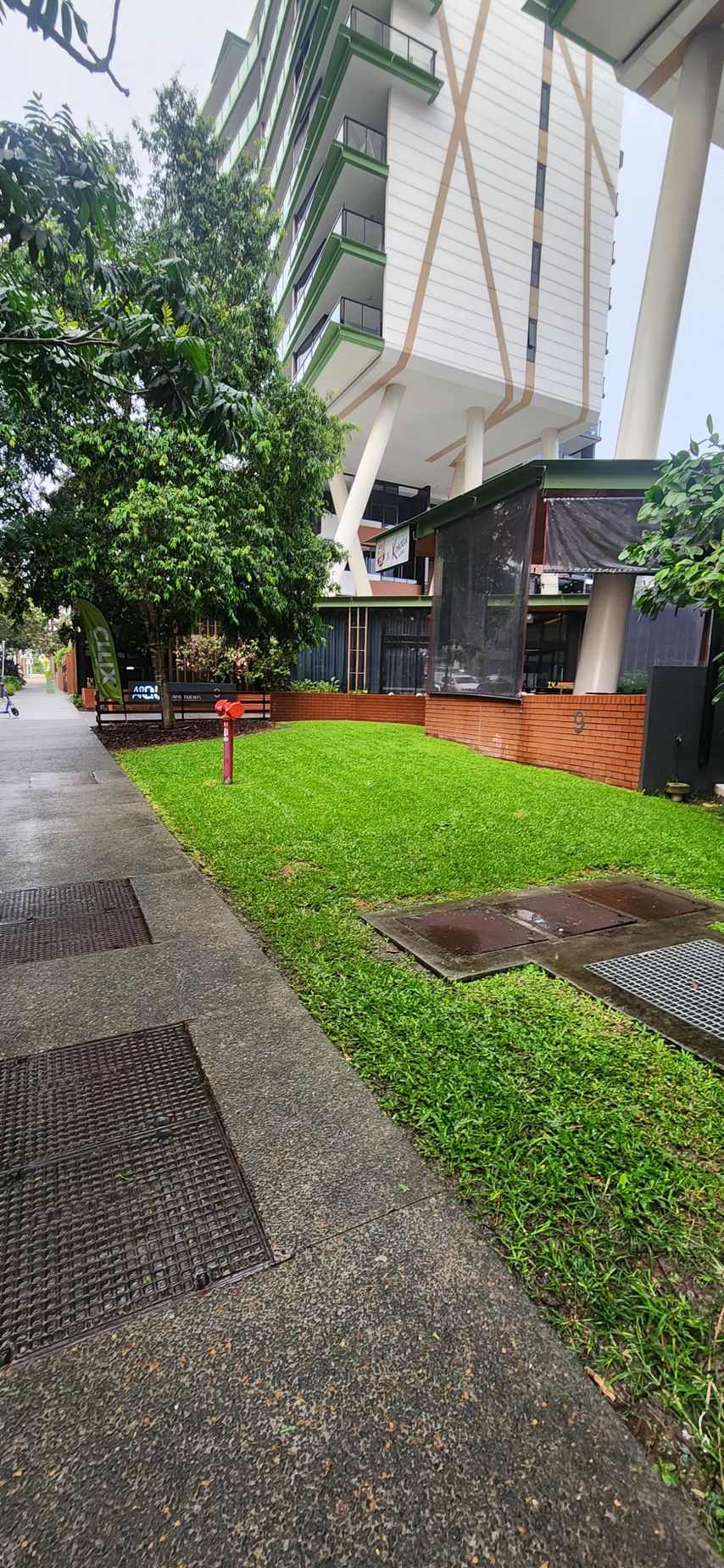 Covered walkway beside a green lawn and trees, with a white building and wet pavement in the foreground.