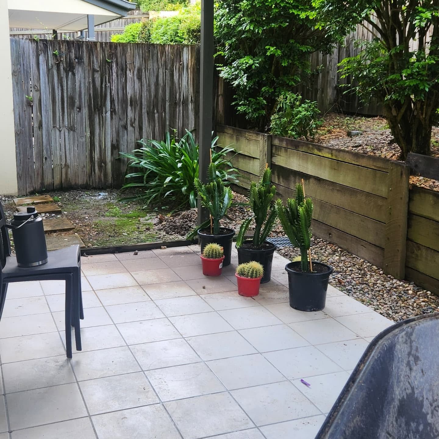 Patio with potted plants beside a wooden fence and garden bed, with a chair and tiled floor.