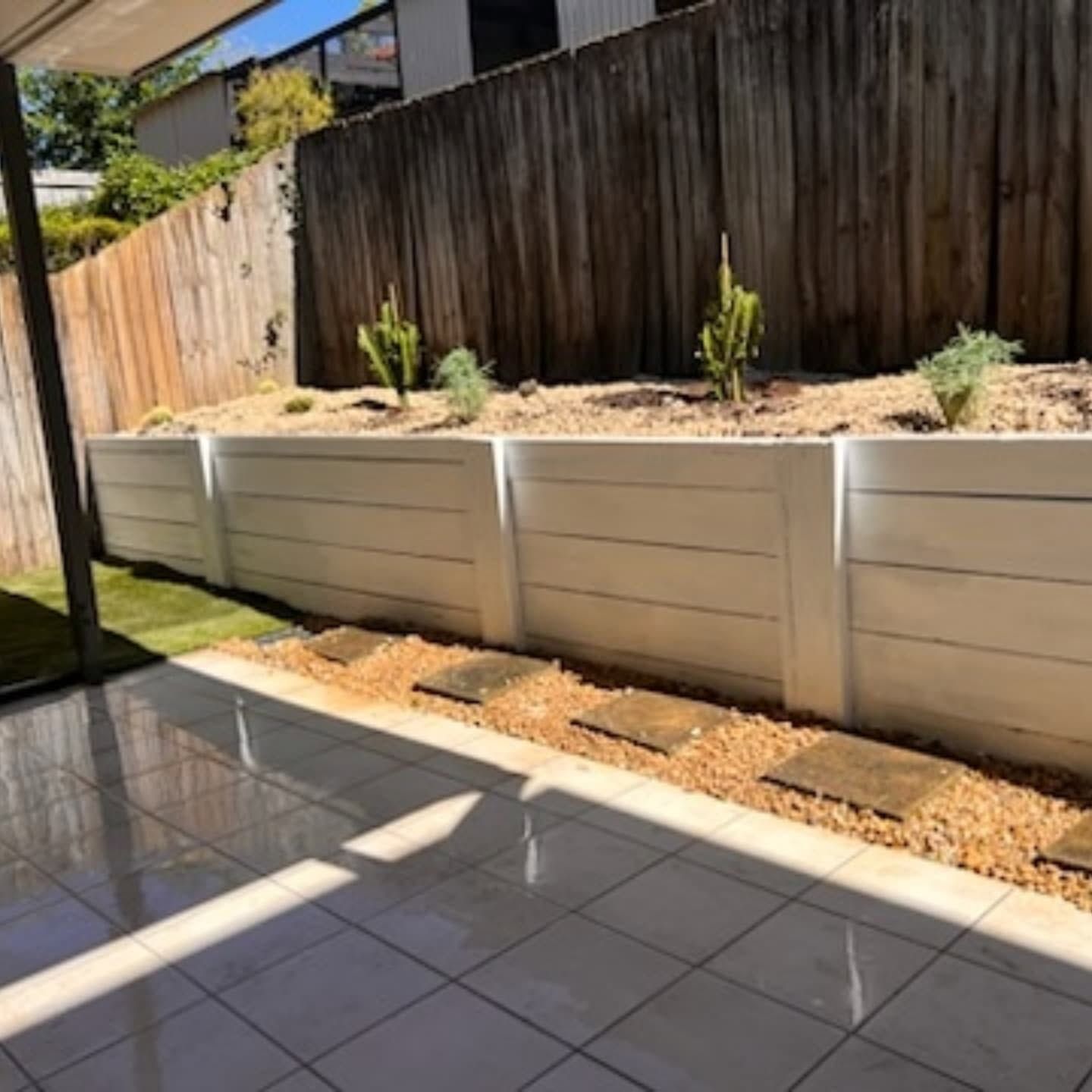 Sunlit patio beside a raised white planter wall and wooden fence with young plants.