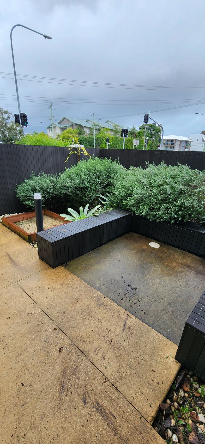 Rainy suburban sidewalk with wet pavement, low black retaining wall, shrubs, and cloudy sky