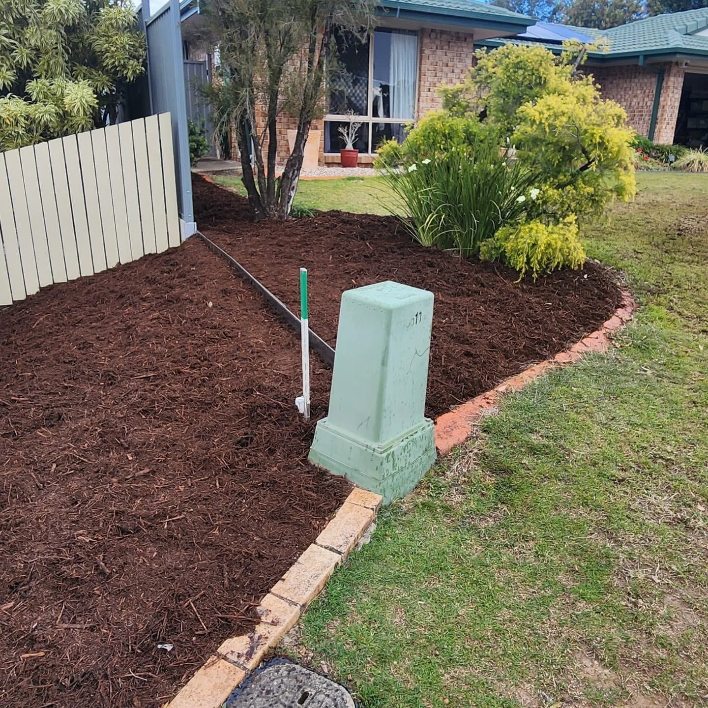 Front yard garden bed with dark mulch, small shrubs, a brick border, and a green utility box near a house