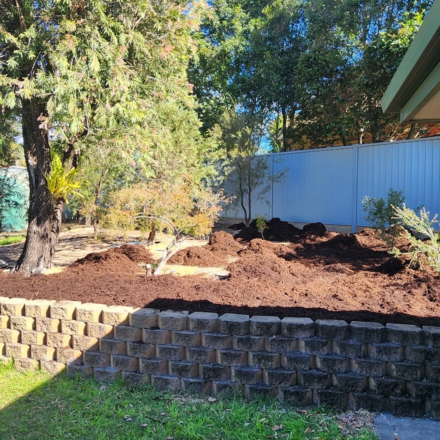 Landscaped yard with new mulch bed, small shrubs, retaining wall, and a white fence in sunlight