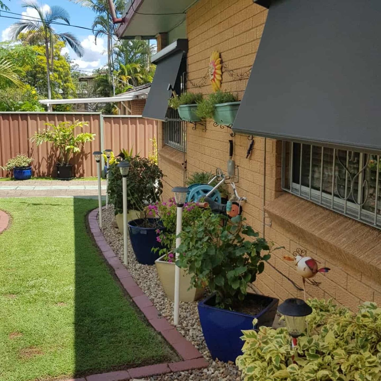 Garden path beside brick house with potted plants, green lawn, and a small bird ornament.