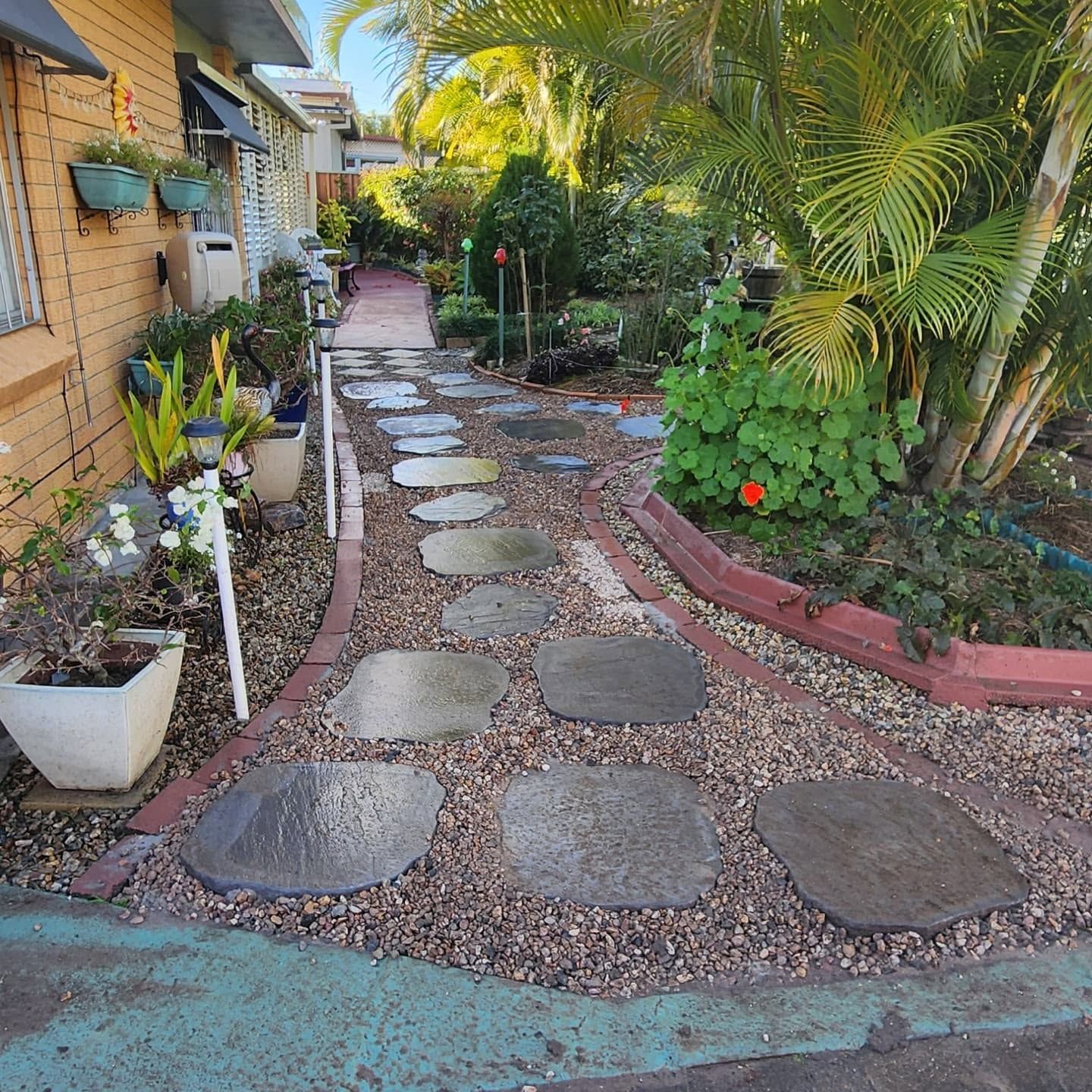 Stone stepping path through a narrow garden beside a house, bordered by potted plants and palm trees.