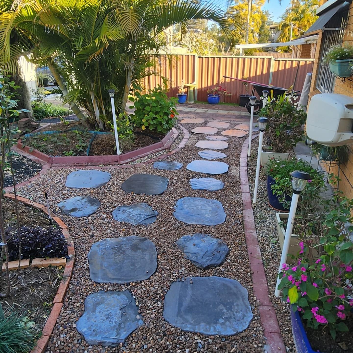 Stone stepping path through a tropical garden toward a wooden fence and patio area.