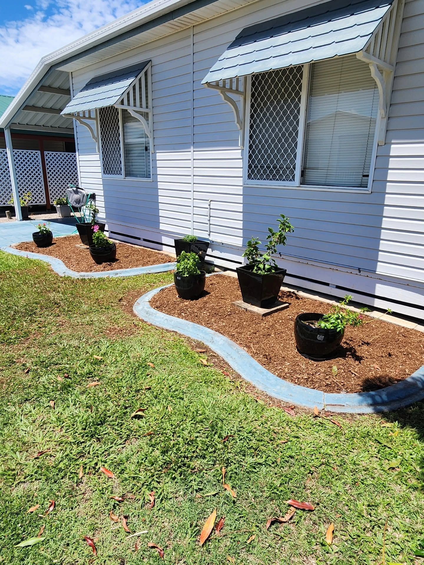White house exterior with striped awnings, potted plants, and a curved blue-edged flower bed along the lawn