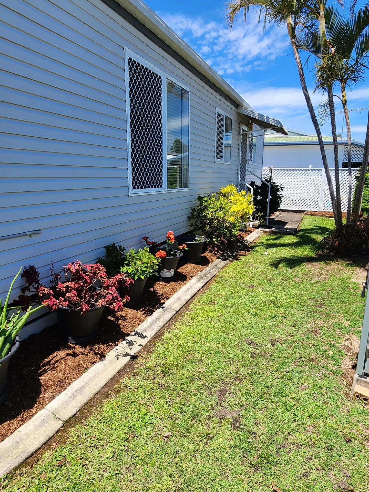 Side yard beside a white house with flower beds, green lawn, and a fence under sunny skies