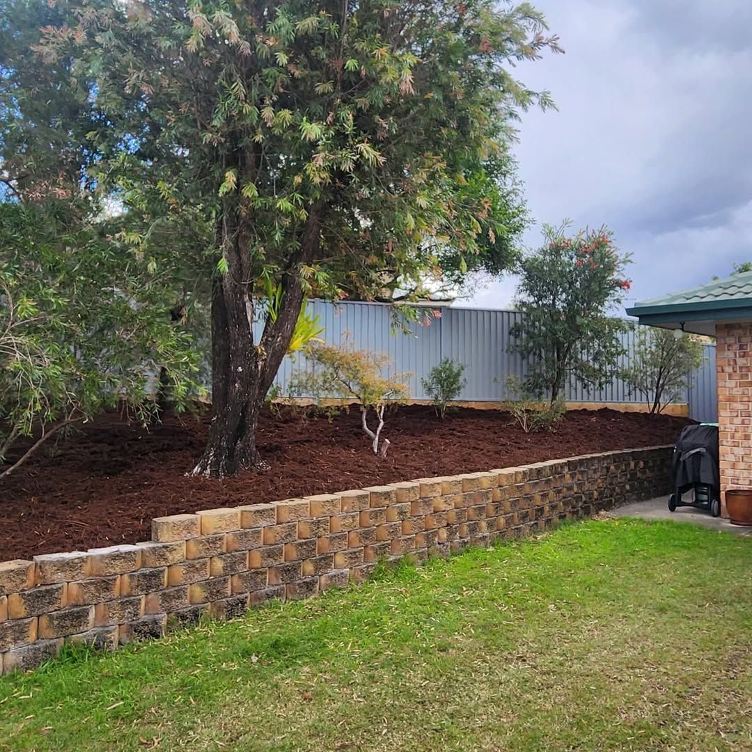 Landscaped backyard with a stone retaining wall, trees, grass, and a corrugated metal fence under cloudy sky