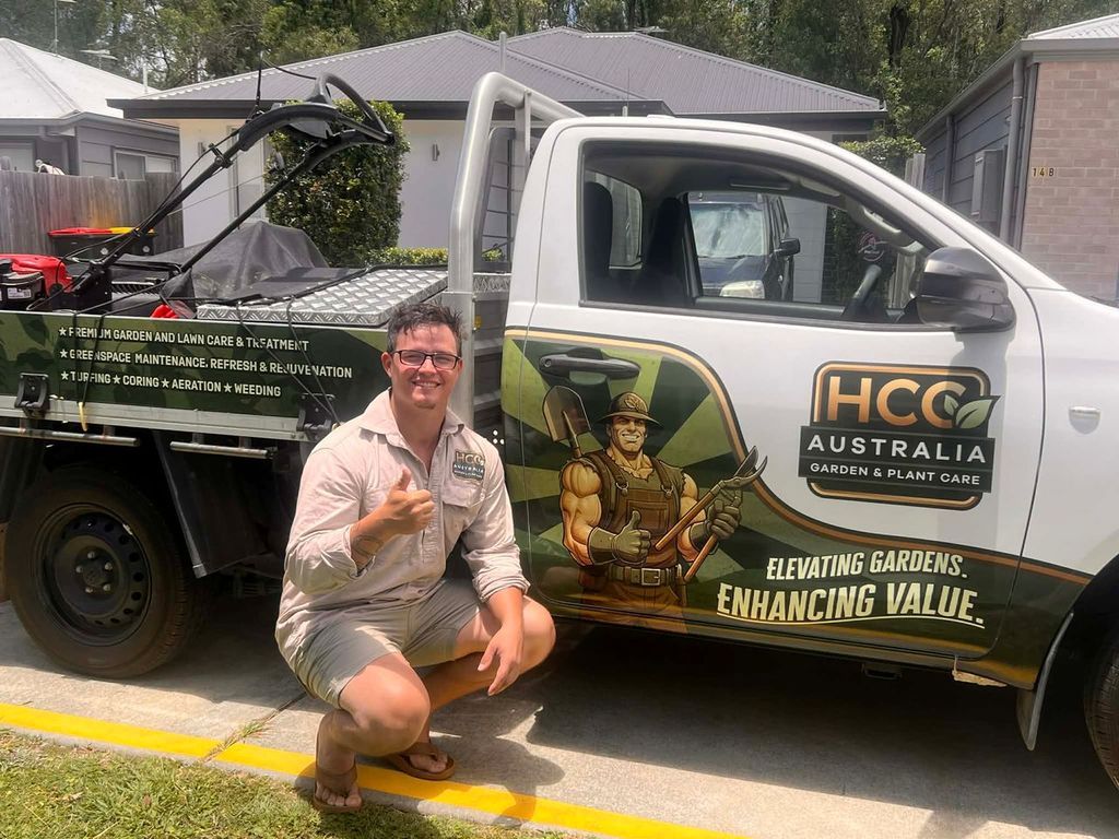 Man kneeling beside a white HCC Australia work ute with landscaping logo in a suburban driveway