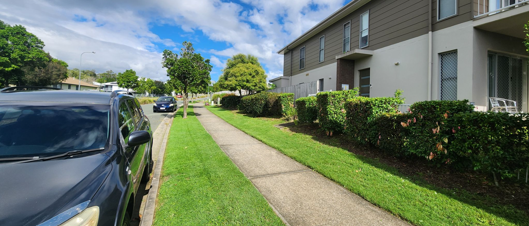 Apartment sidewalk beside parked cars and hedges on a sunny residential street
