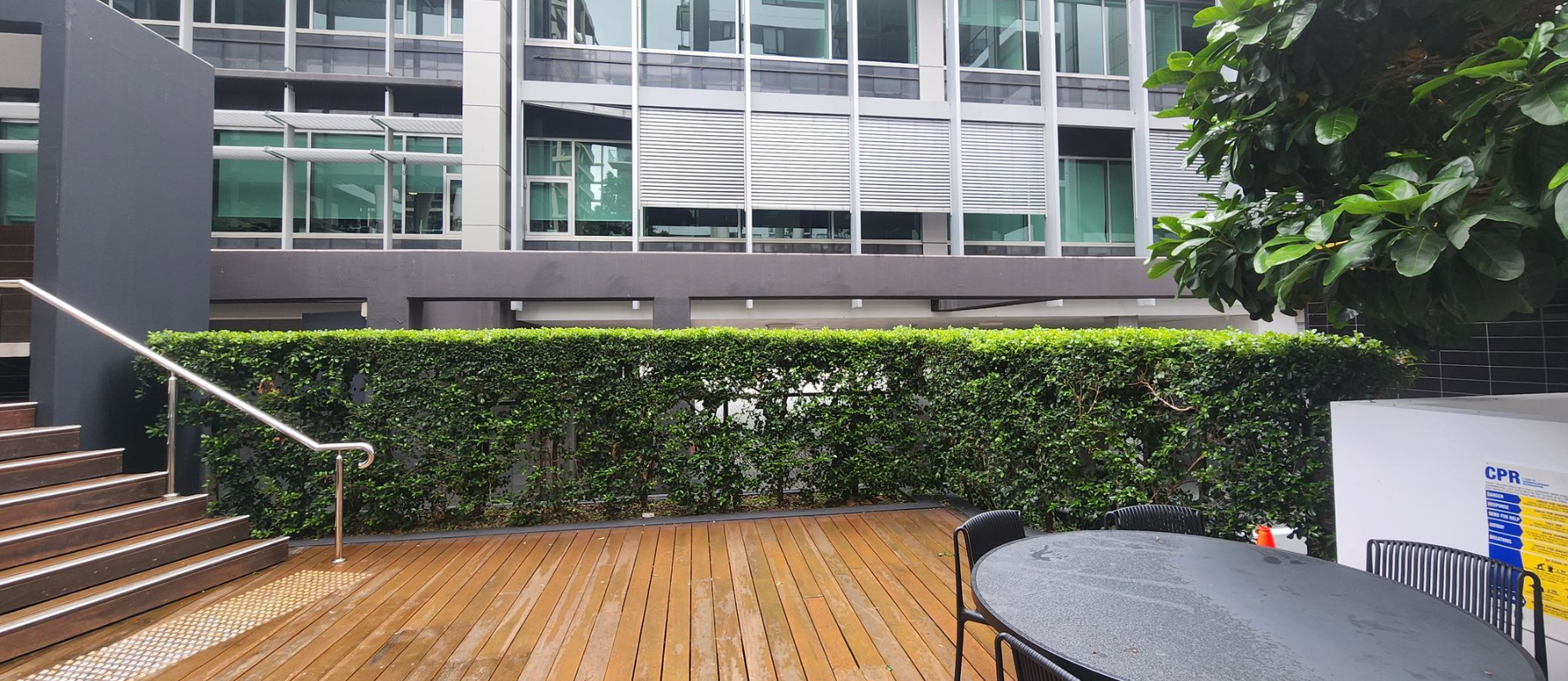 Wooden deck beside modern building with glass windows, hedge, stairs, and a round table under trees