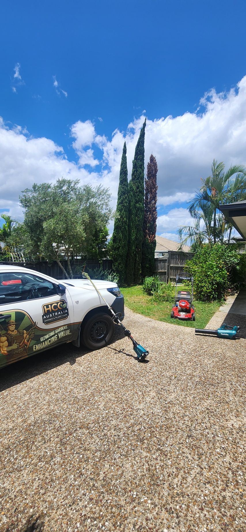 Sunny driveway with a white SUV, gravel yard, tall cypress trees, and garden tools under a blue sky.