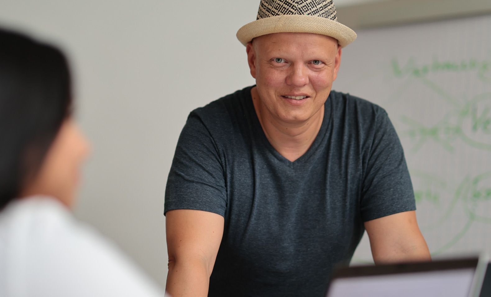 Ein Mann mit Strohhut lächelt eine Person außerhalb des Bildes an, die in einem hellen Raum neben einer weißen Tafel steht.
