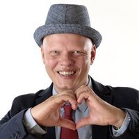 Mann mit rasiertem Kopf, Hut und Anzug, formt mit den Händen ein Herz und lächelt.