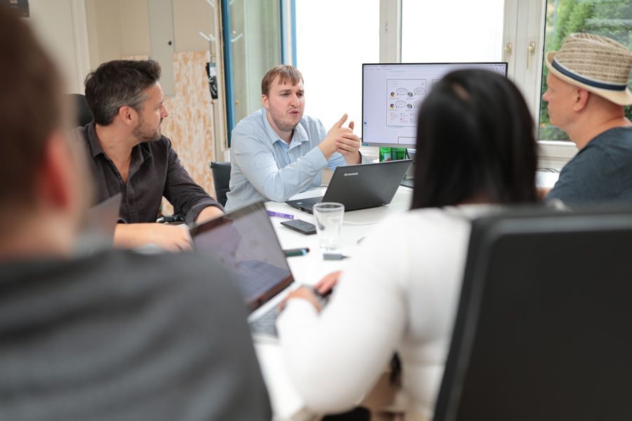 Personen in einer Besprechung, einer spricht am Tisch mit Laptops und einem Monitor, auf dem ein Diagramm angezeigt wird.