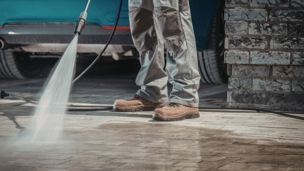 Person pressure washing a concrete driveway beside a brick wall and parked car