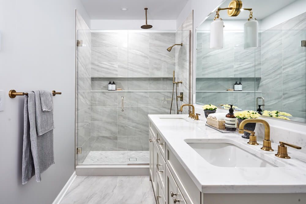 AD Construction NJ - Modern bathroom with marble shower, double sink with gold fixtures, and grey towels.