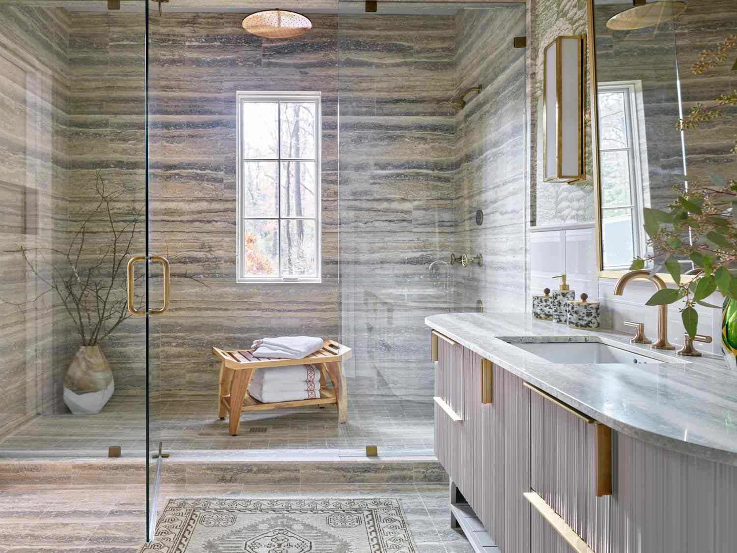 AD Construction NJ - Modern bathroom with stone walls, glass shower, vanity, and window. Beige, gold, and white tones.