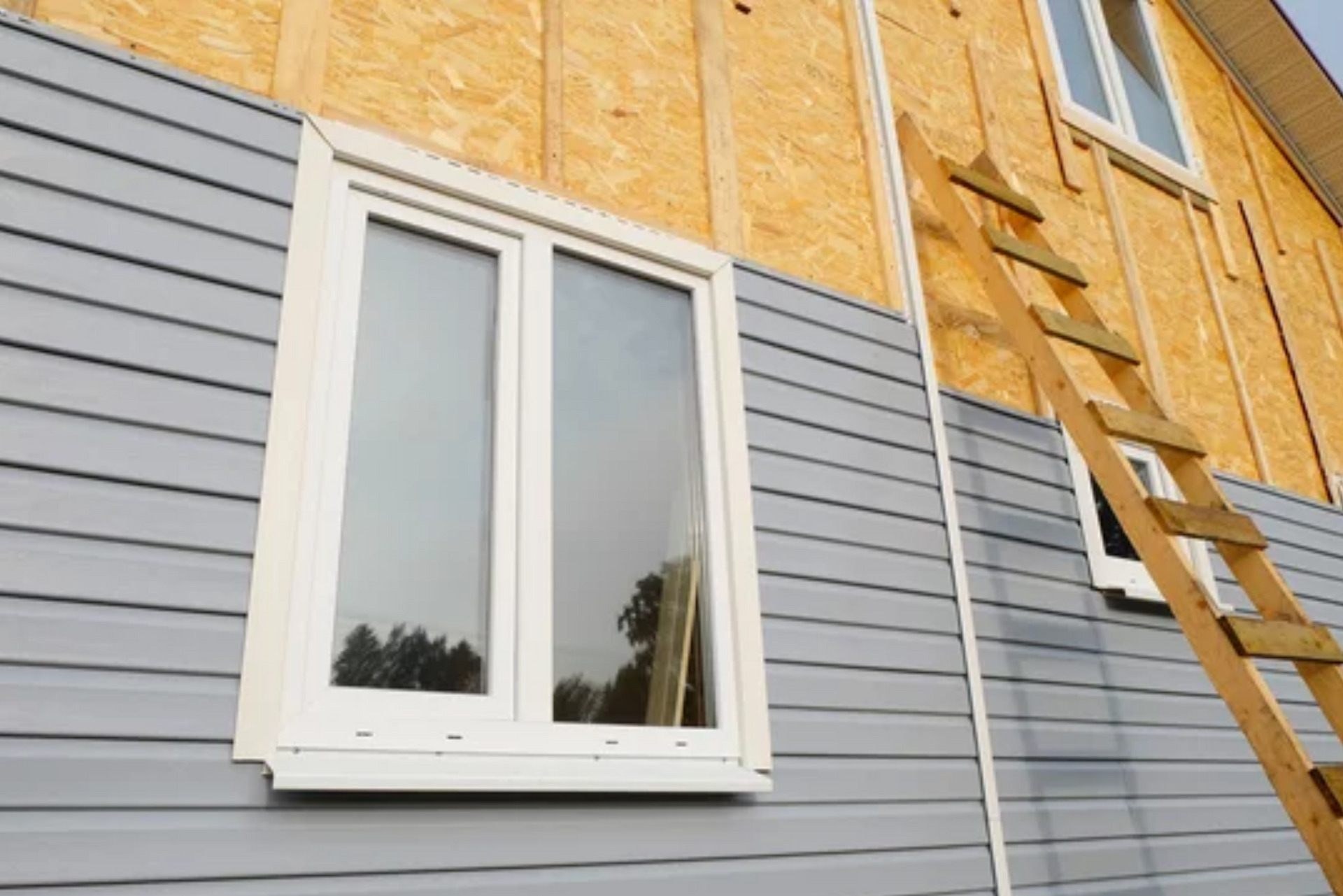 AD Construction NJ - House under construction with gray siding, a white-framed window, and a wooden ladder.