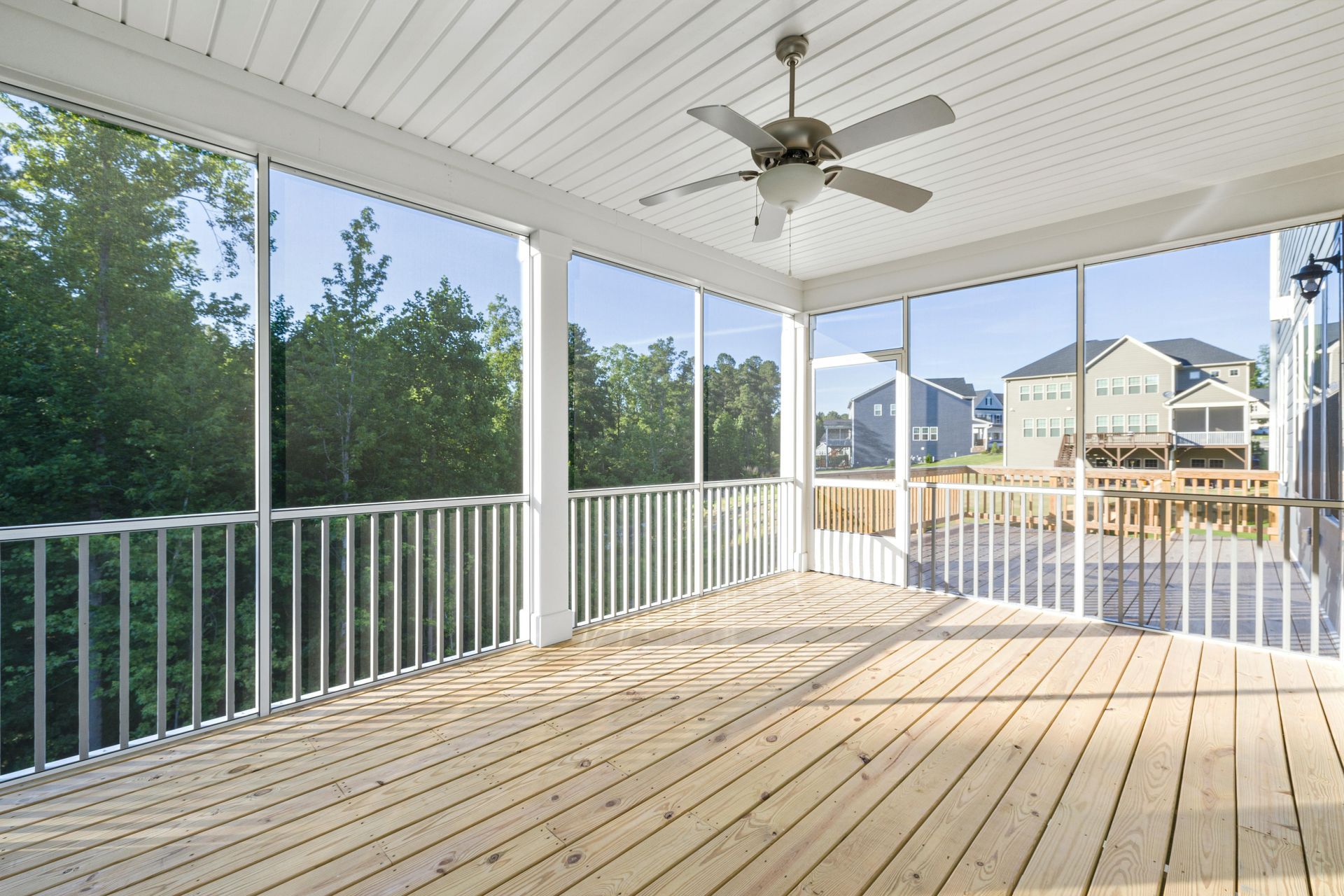 AD Construction NJ - Screened porch with wooden floor, white railing, and ceiling fan; view of trees and houses.