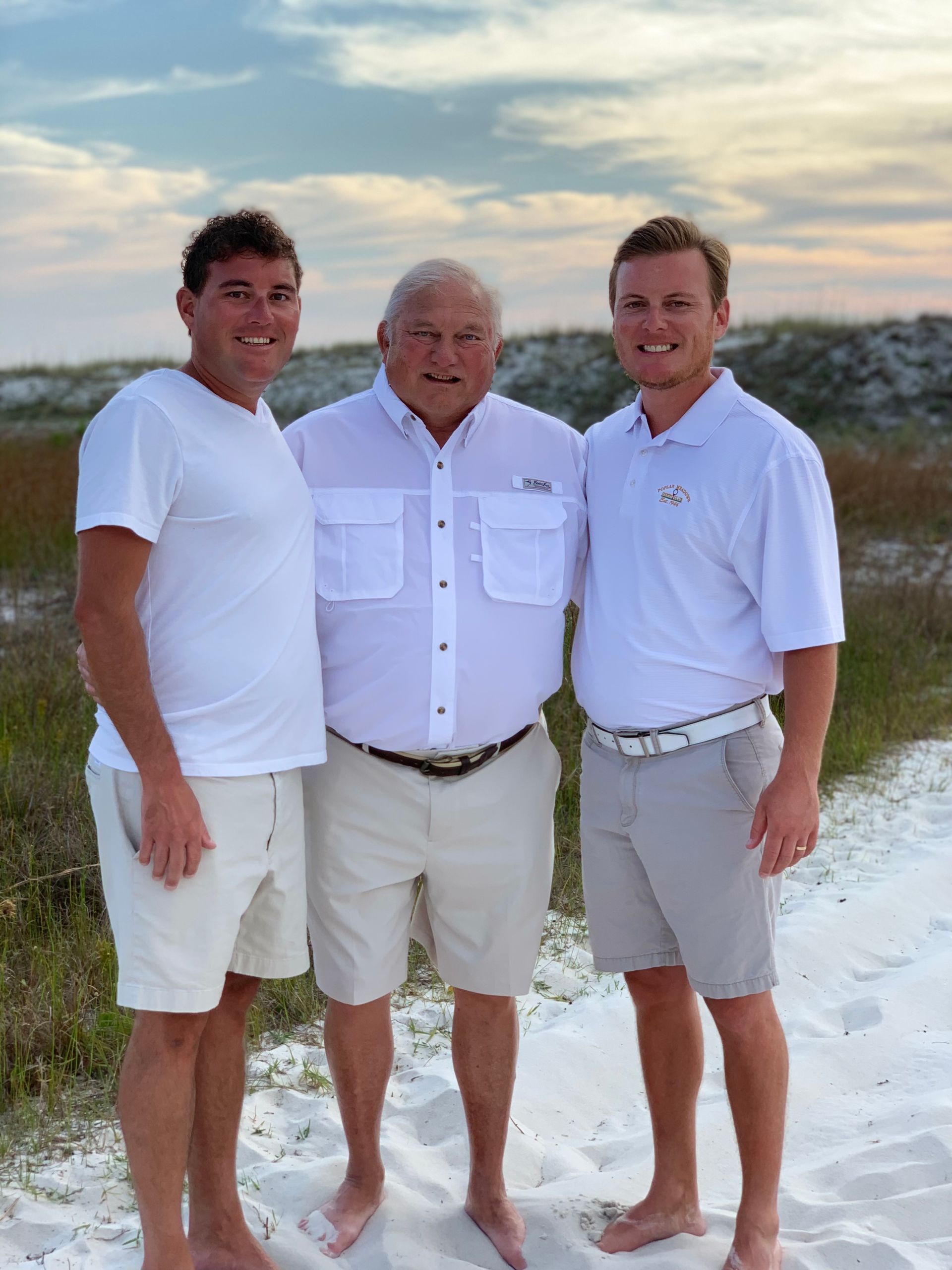 Three men standing barefoot on a beach at sunset, smiling and posing in white shirts and shorts