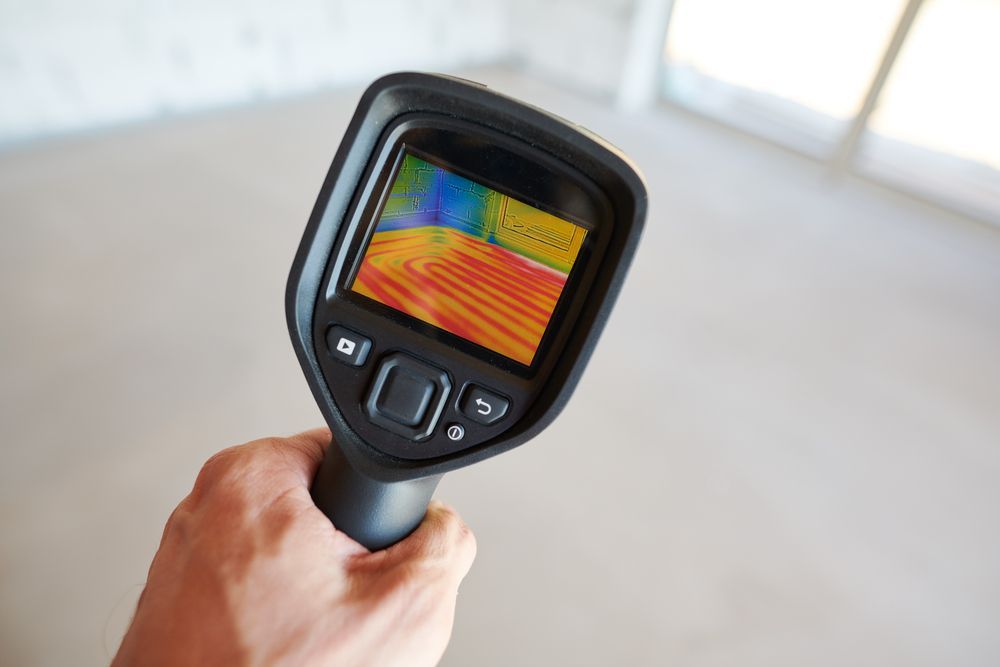 Main tenant une caméra thermique, montrant une carte thermique sur un sol, potentiellement pour l'inspection d'un bâtiment.
