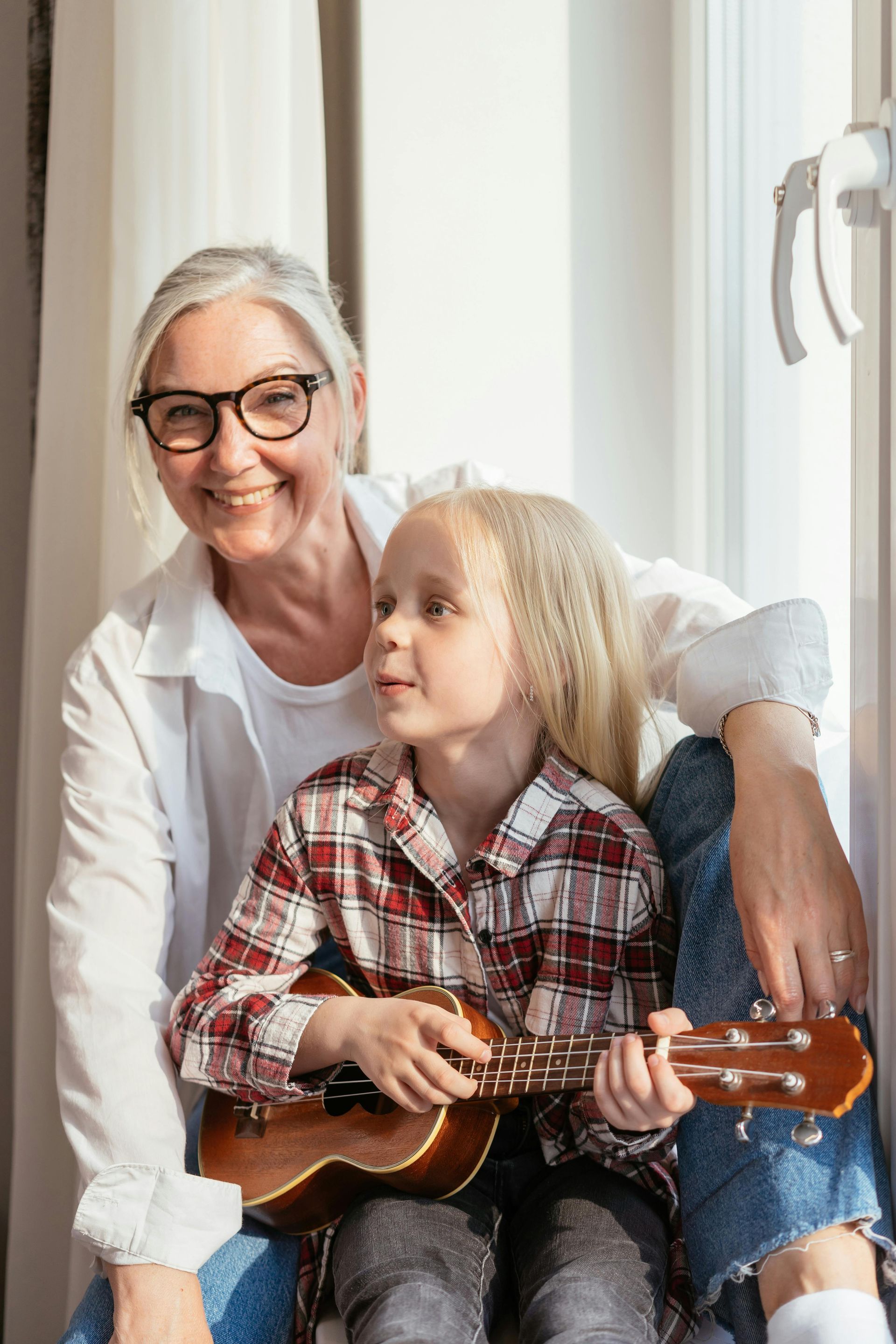 An elderly woman is sitting next to a little girl playing a ukulele.