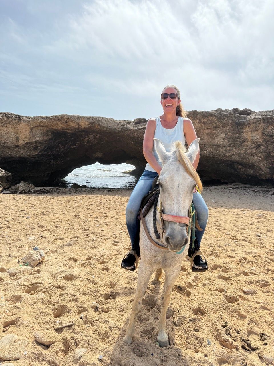 Person riding a white horse on a sandy beach near a rocky arch in Aruba